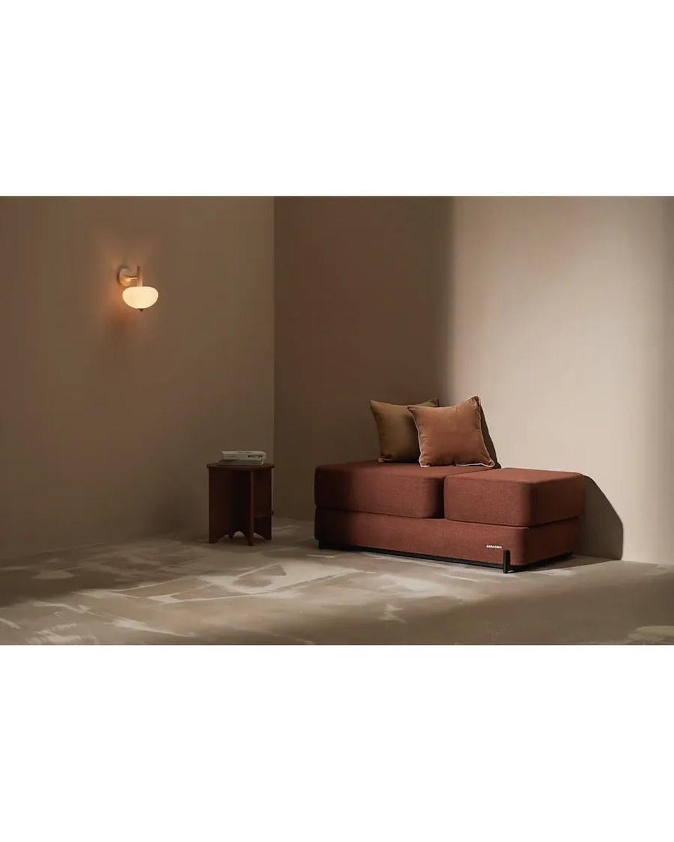 The CERAGEM S4 lounger: a low rust-red upholstered lounger with two cushions, in the corner of a softly lit beige room.
