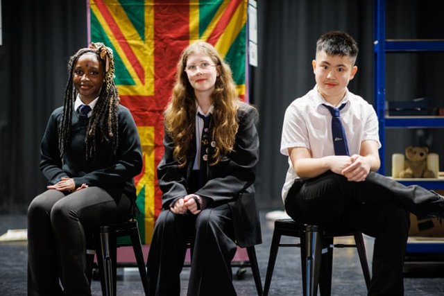 Three secondary-school students in uniform sit on stools on a stage, a red, green and yellow flag behind them.