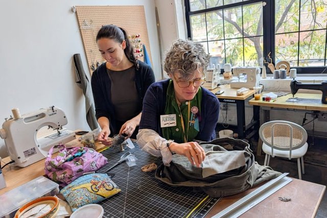 Two volunteers work on a bag at a table crowded with sewing machines and fabric in a bright workshop.
