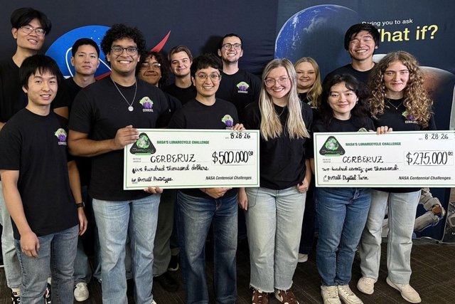 The CERBERUZ team, about 18 students in matching black T-shirts, hold up two oversized prize cheques in front of a space-themed backdrop.
