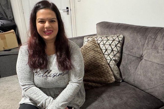 Neelam Heera-Shergill, smiling, sits on a grey sofa beside patterned cushions.
