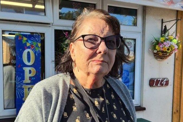 Christine Gardner, in glasses and a grey cardigan, stands outside a building decorated with bunting.