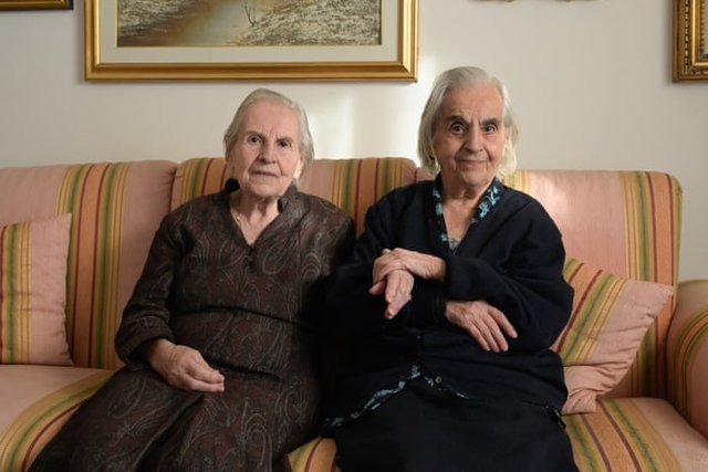The sisters Mariangela and Giovanna Marras, white-haired and in dark clothes, sit side by side on a striped sofa at home.
